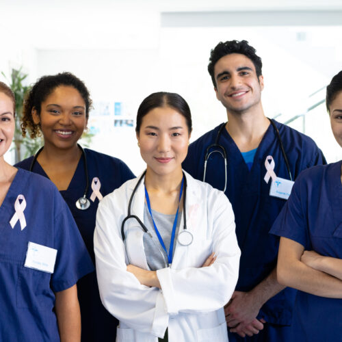 Portrait of diverse group of healthcare workers wearing cancer ribbons smiling in hospital corridor. Hospital, medical and healthcare services.