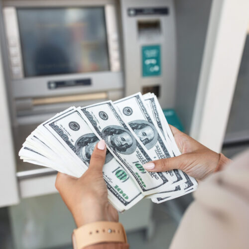 Young arab woman in hijab withdraws money, counts dollars near atm with blank screen. Lady holding cash banknotes outdoor. Finance, transactions, credit card, winning money, currency transfer concept